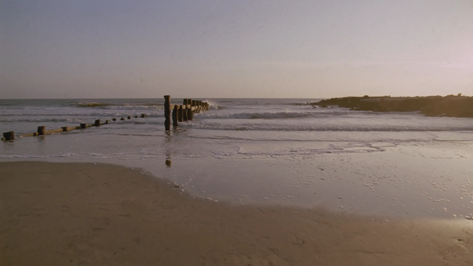 Gentle waves rolling onto a wide sandy beach with wooden groynes at golden hour