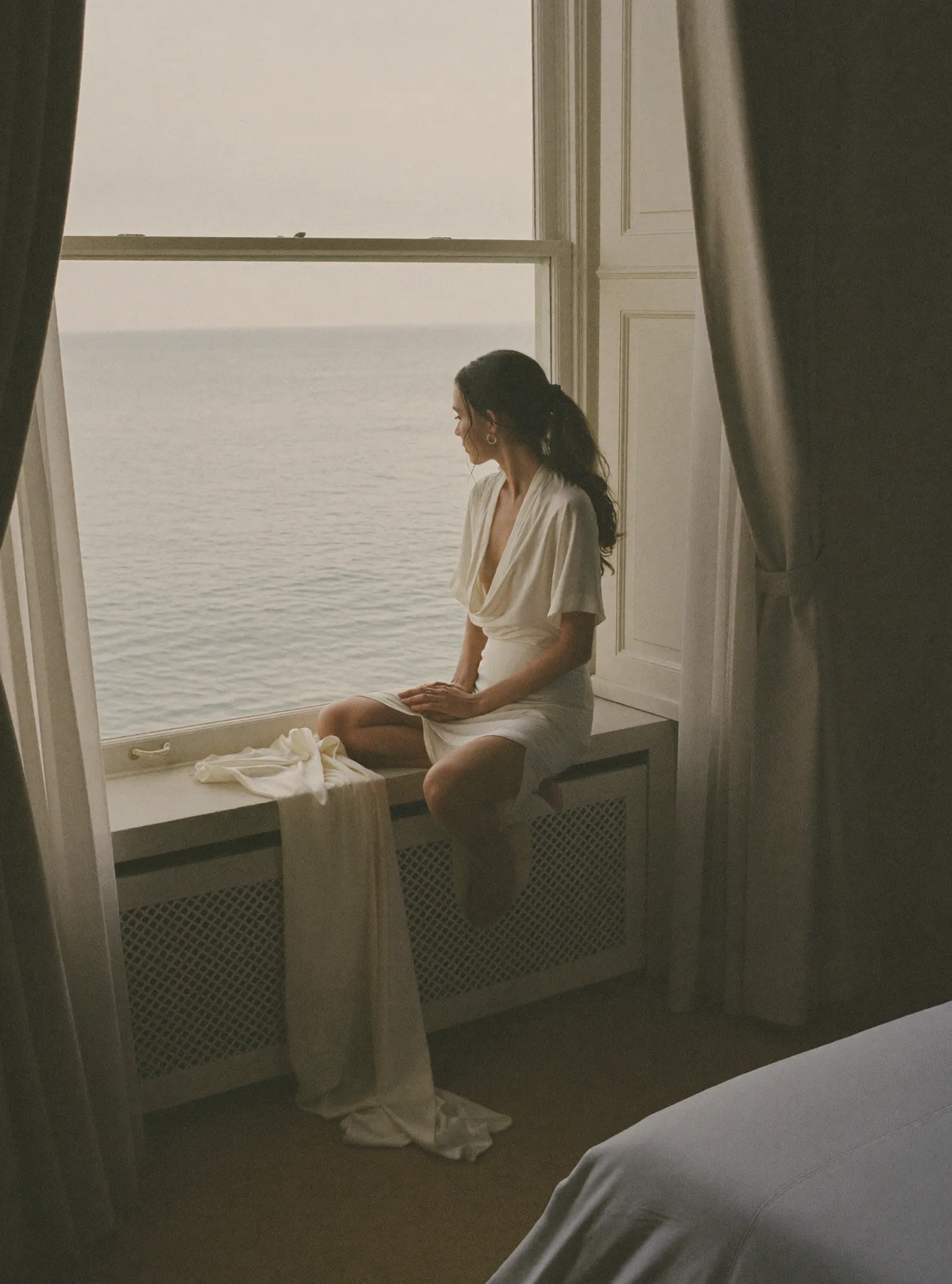 A bride on a window seat looking out to sea on the morning of her wedding